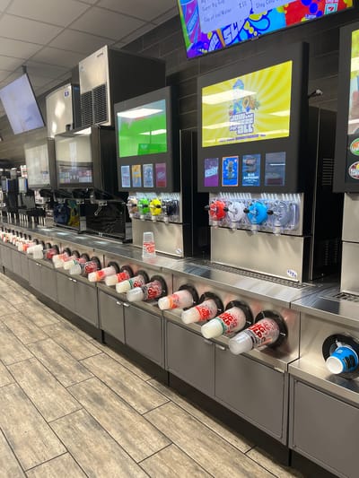 The image depicts a beverage station in what appears to be a convenience store or gas station in Snyderville, Utah, United States of America. The station features a long, stainless steel counter with multiple drink dispensers. In the foreground, several clear plastic cups, labeled "BIG GULP," are inserted into circular openings in the counter, ready for filling. Behind these, a row of soda fountains with multiple spouts is visible, along with what seem to be touchscreen displays for selecting beverages. Further back, there are additional beverage dispensers and large, silver ice machines. The environment has a utilitarian feel, with tiled flooring and dark brick-patterned walls. A large television screen is mounted high on the wall, displaying an advertisement. No people are visible in the scene.