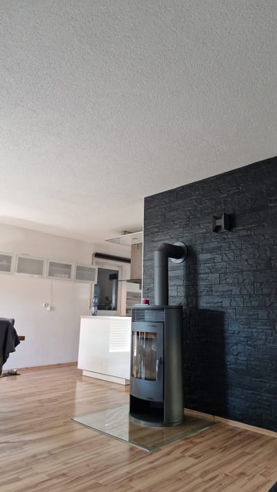 The image depicts the interior of a room, likely a living area and kitchen in a residential property located in Ichenhausen, Germany. The foreground features a modern, dark gray wood-burning stove situated on a clear glass hearth plate, with its flue pipe extending upwards and to the left. The flooring is light-colored laminate, reflecting light. To the left, partially visible, is a dining table with a dark cloth draped over it.

In the mid-ground, the space transitions into a kitchen area with white cabinetry and a countertop. A bottle of water and a small red glass are visible on the countertop. Above the kitchen counter, there are several white wall-mounted cabinets with frosted glass doors. A window with blinds is also visible in the kitchen area.

The background reveals a ceiling with a textured finish. A prominent feature of the room is a wall covered in dark gray, textured stone-like tiles, behind the wood-burning stove. A small, dark vent-like object is mounted on this tiled wall, to the right of the flue pipe. The overall lighting suggests daylight, with sunlight casting shadows. No individuals are present in the image.
