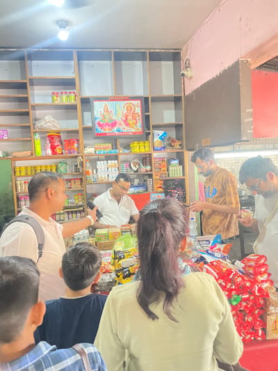Here is a description of the image:

The image depicts the interior of a small shop, seemingly located in Katauhar Kalan, India. Patrons are gathered near shelves stocked with various items, including snacks and household products. A poster of Hindu deities hangs on the shelf. Customers are seen interacting with the shop's merchandise and personnel. A security camera is mounted near the ceiling.