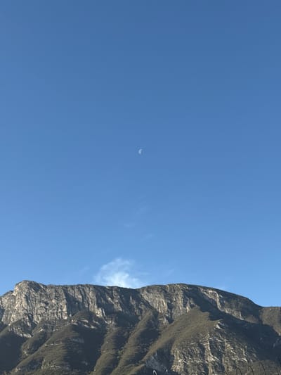The image displays a vast outdoor scene located in Ciudad Santa Catarina, Mexico, featuring a prominent mountain range under a clear blue sky. The mountain, likely part of the Sierra Madre Oriental, occupies the lower half of the frame, presenting steep, rugged terrain. Its upper sections consist of grey and white exposed rock formations with vertical striations, indicative of limestone cliffs. The lower slopes are densely covered in dark green scrub or low vegetation. In the upper right quadrant of the deep blue sky, a pale, waxing crescent moon is visible. A few faint, wispy white clouds are present near the horizon line, just above the mountain peaks. No individuals, artificial structures beyond the landscape, or specific actions or interactions are depicted. The lighting suggests daytime conditions.