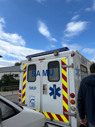 The image shows the rear of a white ambulance with red and yellow striped markings. The word "SAMU" is written in blue on the back of the ambulance. There is a blue star of life on the ambulance. The ambulance is parked on a paved road in front of a building with a green roof. A man is standing behind the ambulance. The image was taken in Dinard, France, which is a town on the coast of Brittany, known for its beaches and coastal walks. The coordinates provided locate the scene in a residential area near the town center.