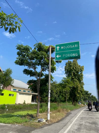 The image shows a road sign in Kecamatan Tulangan, Indonesia, located at coordinates -7.475 latitude and 112.634 longitude. The sign indicates two directions: "Mojosari" to the right, and "Porong" to the left.  The sign is mounted on a metal pole, and the background consists of a paved road, green grass, a small canal, and lush greenery.  The road is straight and runs horizontally across the image. A small, single-story building is located behind the sign, with a bright green wall. Three motorbikes are driving on the paved road in the distance, heading towards the right.  The sky is blue with white clouds.  The trees in the background are all green.