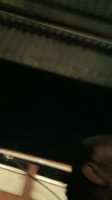 An extreme close-up captures a dark, obscured scene. To the left, fragments of a window and a hint of human features are visible. The central area plunges into near-total darkness. To the right, a curtain or drapery displays varying shades of green, marked by vertical folds and the suggestion of intricate stitching. The image's overall dim lighting and tight framing contribute to an air of mystery.