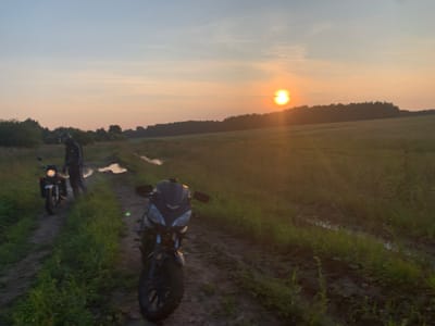 The image depicts two motorcycles parked on a dirt track in a rural setting during sunset. The sky is a gradient of soft blues and oranges, with the sun low on the horizon, casting a warm glow. The track is muddy and has puddles of water, suggesting recent rain. Lush green grass and vegetation flank the track, and a line of trees forms the distant background.

In the foreground, a dark-colored motorcycle with a sleek design is parked, facing towards the right of the frame. Its headlights are off, and the front wheel is turned slightly. Further back on the left, another motorcycle, lighter in color and possibly an adventure bike, is parked. A person wearing a helmet and dark clothing is standing next to this motorcycle, appearing to be taking a break.

The overall atmosphere is peaceful and serene, capturing a moment of pause during a motorcycle ride in the countryside of Verkhnyaya Osh'ya, Russia. The lighting and natural elements suggest a natural landscape devoid of urban infrastructure.