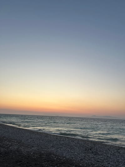 The image captures a serene coastal scene at what appears to be sunset or sunrise in Rhodes, Greece. The foreground is a dark, pebbled beach. The middle ground is occupied by the Aegean Sea, its surface rippled by gentle waves that break upon the shore. In the distance, a hazy silhouette of islands can be observed. The sky transitions from a pale blue at the top to a soft orange and pink hue near the horizon, suggesting the low angle of the sun. There are no people or man-made structures visible on the beach itself. A faint shape of a ship is visible on the horizon.