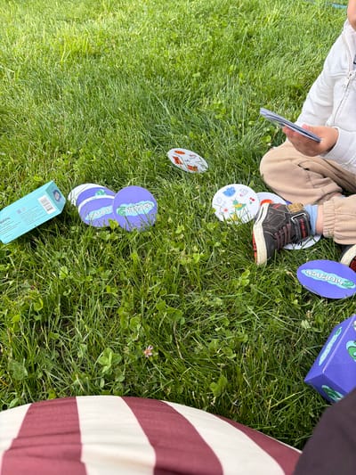 A child sits on a patch of green grass strewn with clover, likely in an outdoor space. The child is wearing tan pants, a white jacket, and black shoes with red laces. They hold a stack of cards in their hand. Several circular cards with cartoon images are scattered around, some lying under the child's shoe. A blue box and a purple box are also on the grass. The presence of a striped object in the frame is evident. The location is potentially in Chkalov, Armenia.