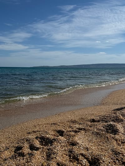 The image depicts a beach scene in Береговой сельский совет, Ukraine. The view is oriented so that the sky and the beach are on the left and right, respectively. The sky is partly cloudy with blue patches. The sea occupies the middle portion, with gentle waves lapping at the sandy shore. The sand is coarse and textured, with visible footprints. In the distance, there is a suggestion of land meeting the horizon, indicating the opposite shore or a nearby elevation.