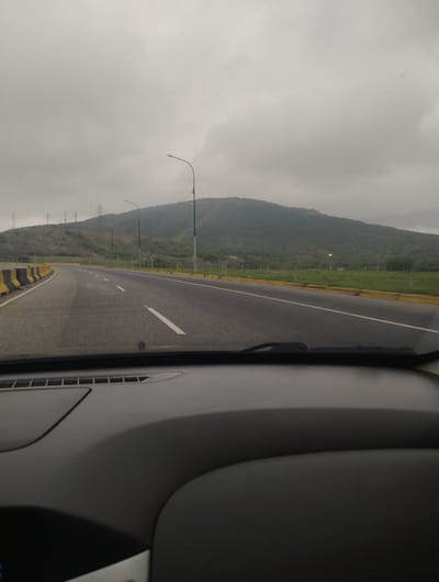 The image depicts a view from inside a vehicle, likely a car, driving on a highway in Barquisimeto, Venezuela. The road is asphalt, with white lane markings and yellow and black safety barriers on either side. To the right of the road, there's a grassy field area. Beyond the field, a large, green, gently sloping hill or mountain dominates the landscape. Power lines and lampposts line the edge of the road and hillside. The sky is overcast and grey, suggesting cloudy weather. The dashboard of the vehicle is visible in the foreground, filling the lower portion of the image.