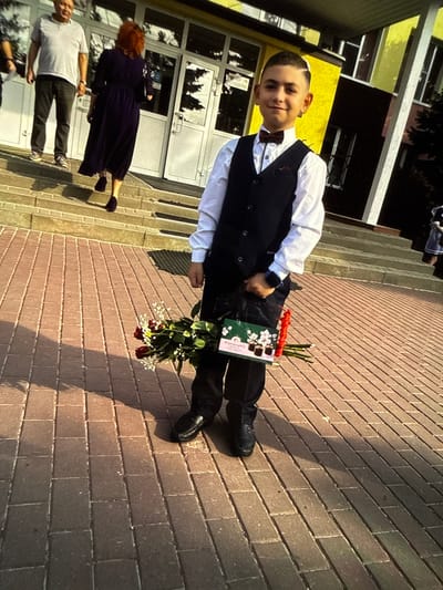 A young boy in a formal outfit stands on a brick walkway in front of a building. He is wearing a white shirt, a dark vest, and a red bow tie. He holds a bouquet of red roses and a small green box. His left hand is on the box, and his right hand is behind his back. The building behind him has yellow and dark brown accents and large glass doors. Two adults, a man and a woman, are walking up steps to the left of the boy. The man wears a light gray t-shirt and dark pants, and the woman wears a dark dress. The scene is lit by sunlight, casting shadows on the brick walkway. The location is identified as Artik, Armenia.