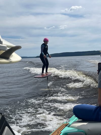 A person in a wetsuit and pink helmet is wakefoiling on a lake, riding behind a boat that is not fully visible. The person is standing on a foilboard, which is lifting them out of the water. The water is choppy and creating a wake. The scene is set in a natural environment with a tree line visible in the distance, suggesting a lakeside location. The sky is partly cloudy. The time of day appears to be daytime. A portion of the boat's interior is visible on the right side of the frame, with light-colored seating and wooden accents. There is also a glimpse of another wakeboard or surf-style board in the foreground, also with a fin. The person on the foilboard appears focused and engaged in the activity. Some text is visible on the person's wetsuit, appearing to be a number "76".