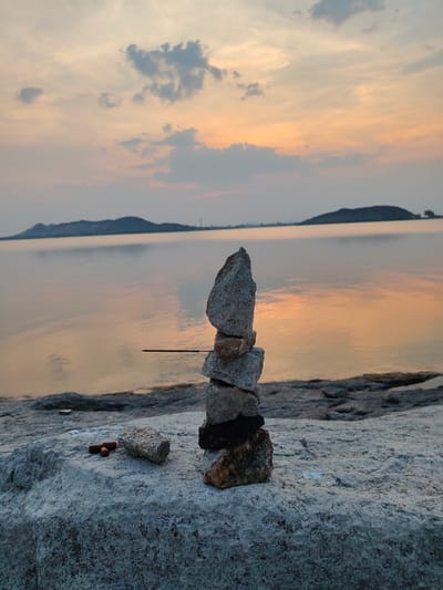The image depicts a rock cairn on a rocky shoreline in Kammavaripalli, India, with a serene lake and distant hills under a sunset sky in the background. The cairn consists of several stacked stones of varying sizes and colors, balanced on a large, flat, weathered rock. A thin stick is wedged between two of the rocks in the stack. Near the base of the cairn, on the flat rock, are two small, dark brown cylindrical objects and another small rock. The lake reflects the orange and gray hues of the sunset. The hills in the distance are silhouetted against the horizon. The sky contains a mix of gray and orange clouds. The scene is still and quiet, suggesting a peaceful moment at dusk.