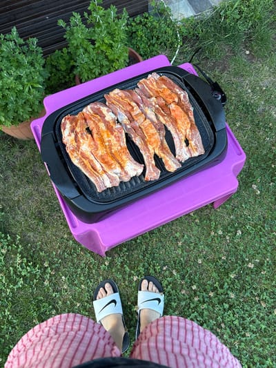 This image captures an outdoor grilling setup in Polena, Bulgaria. A black electric grill, loaded with approximately seven to eight pieces of raw, seasoned meat (likely pork ribs) exhibiting an orange-red spice coating, rests on a vibrant purple plastic stool or low table. The grill's integrated control unit, marked "THERMOSTAT CONTROL," is visible on the right, with an electrical cord extending towards the background. The ground is covered in green grass interspersed with numerous small white clover flowers. In the background to the left, two potted plants with dense green foliage are visible, along with a dark wooden fence or structure. The lower portion of the frame shows the bare feet of an individual wearing black and white Nike slide sandals, with a segment of pink and white striped shorts or trousers also visible, indicating a first-person viewpoint of the scene.