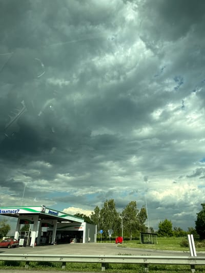 The image displays a gas station with a distinctive green and white canopy, labeled "ADVANTAGE 7" and "ADVANTAGE." Several fuel pumps are visible, with a red pickup truck parked at one of them. To the right of the gas station, a road is bordered by a metal guardrail and a grassy embankment. In the background, lush green trees and foliage suggest a rural or suburban setting. Above, a dramatic sky dominates the scene, filled with heavy, dark clouds interspersed with patches of bright, diffused sunlight. This creates a sense of impending weather change, possibly rain, and contributes to a somewhat moody atmosphere. The time of day appears to be daytime, given the overall illumination, but the cloud cover makes it difficult to determine precisely. No people are clearly visible in the frame, but the presence of the parked truck implies some activity at the gas station. The overall impression is one of a transient moment, captured from a moving vehicle, with the focus on the interplay between the man-made structure of the gas station and the powerful natural forces of the sky. The location context provided indicates Mytnytsia, Ukraine.