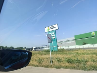 The image captures a scene on a sunny day in Ötegen Batyr, Kazakhstan. A tall, green sign with the text "Damu Logistics" is prominently displayed, featuring the company's logo. To the right of the sign, a large, green industrial building with a white fence in front of it dominates the background. Several white trucks are parked in a lot in front of the building. The foreground shows a road and dry grass, with the side mirror of a vehicle reflecting the scene. The sky is clear and blue with a few wispy clouds. The overall impression is of an industrial area on a clear day.
