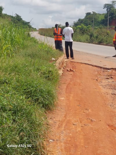 Two individuals are standing on the side of a road. One person, wearing a white shirt and black pants, stands closer to the road. The other person, to the left and slightly behind, is wearing a high-visibility orange vest over dark clothing. The ground beside the road is uneven and consists of reddish-brown dirt. Lush green grass and vegetation grow along the edge of the road and in the foreground. In the background, a tree-covered hillside is visible under a cloudy sky. The scene suggests an outdoor setting, possibly in a rural or developing area. There are no explicit indications of the time of day or weather conditions beyond the overcast sky. The visible text "Galaxy A52s 5G" is a watermark, indicating the device used to capture the image.