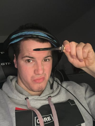 A person with light skin, brown hair, and brown eyes is holding a narrow object, possibly a metal straw or a writing instrument, close to his forehead. He is wearing a headset with a blue accent band around his head. He is also wearing a gray hooded sweatshirt. The background is dark. The image provides insufficient information to confirm the location as Benningen, Germany.
