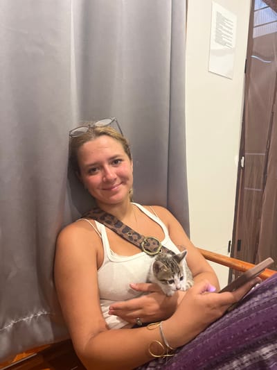 An indoor scene in Baan Nai Wok, Thailand, depicts a female individual holding a kitten while seated. The female, with light hair and clear-framed glasses resting on her head, wears a white tank top, purple patterned pants, and a leopard-print shoulder strap. She displays several bracelets on her left wrist and a ring on her right ring finger. She is smiling and looking directly at the viewer. A small, white and tabby-patterned kitten is cradled in her arms, looking towards the right. In her left hand, she holds a dark-colored smartphone with an illuminated screen. The background features a light grey vertical curtain on the left and an off-white wall on the right. A framed document or paper is affixed to the wall, and a partial view of a brown wooden door or window frame with vertical elements is visible on the far right. A light brown wooden armrest is visible to the female's right.