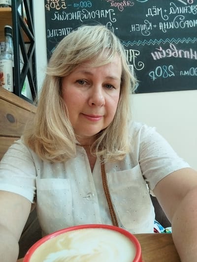A woman with blonde hair is taking a selfie indoors. She is wearing a white blouse and has a cup of coffee in front of her. A blackboard with writing is visible in the background. The image appears to be taken in a cafe or restaurant.