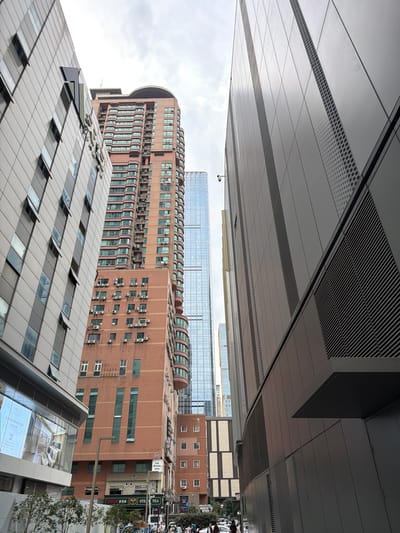 The image shows a street scene in Qinhuai District, China, with a focus on modern architecture. On the left, a multi-story building with a light-colored facade is prominent, featuring numerous windows, some of which are open. To the right of this building is a tall, reddish-brown brick building with many evenly spaced air conditioning units mounted on its exterior. Behind these structures, a very tall, slender glass skyscraper rises into the cloudy sky, reflecting the surrounding buildings and the sky itself. Another modern building with a dark gray, textured facade is situated in the foreground on the right, creating a canyon-like effect with the other buildings. The street level shows a few pedestrians walking, and vehicles, including a white van and other cars, are visible further down the street. Several signs in Chinese are present on the buildings at street level. The overall impression is an urban environment with a mix of older and contemporary architectural styles.