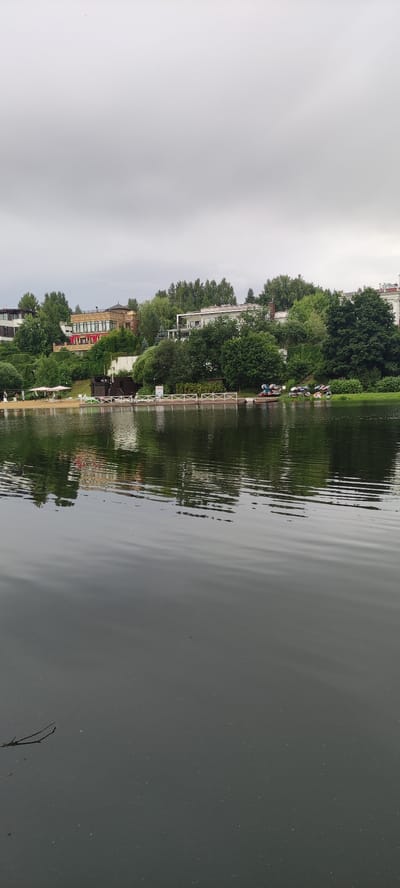 The image depicts a cloudy day scene at a waterfront in Angelovo, Russia. In the foreground, the dark water of a lake or river fills the lower half of the frame, with subtle ripples distorting the reflection of the shore. A small, thin branch floats on the surface in the lower left corner. The middle ground reveals a sandy beach area with several white beach umbrellas and lounge chairs. Adjacent to the beach is a wooden dock with white railings. On the dock and further along the shore, several small boats, some with people near them, are visible, as well as some wheeled carts. The background is dominated by lush green trees and several multi-story buildings, characteristic of a residential or resort area. The buildings have a modern architectural style with various colors and materials. The sky is overcast and light gray, indicating a subdued lighting condition.