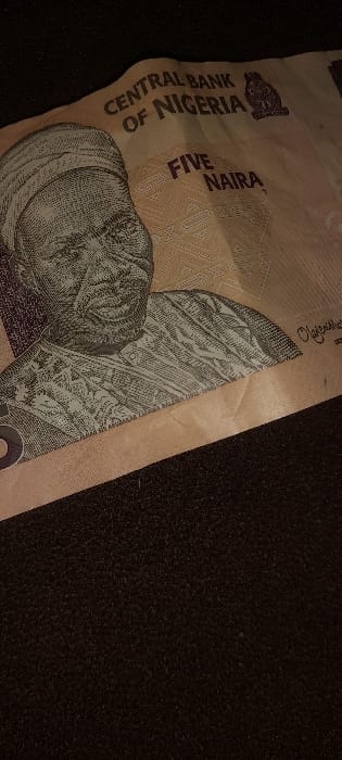 A Nigerian Five Naira banknote, slightly creased, is depicted resting on a dark, textured surface. The banknote features a portrait of an elderly man wearing a traditional cap and attire, with the inscriptions "CENTRAL BANK OF NIGERIA" and "FIVE NAIRA" prominently visible. No individuals or active events are present in the scene. The setting is in Ashipa, Nigeria.