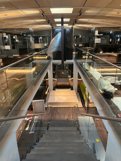 An interior shot looks down a modern wooden staircase with glass railings. The staircase is in a multi-level building, possibly a museum or exhibition space, with the ceiling featuring wooden slats and integrated lighting. Displays behind glass are visible on the upper levels, some covered with protective sheeting. Various objects, including what appear to be large crates, bubble wrap, and black trash bags, are present on the landings and along the sides of the stairwell. The scene suggests a period of setup, renovation, or closure for the space. The image does not provide information to identify the specific city of Cologny, Switzerland.
