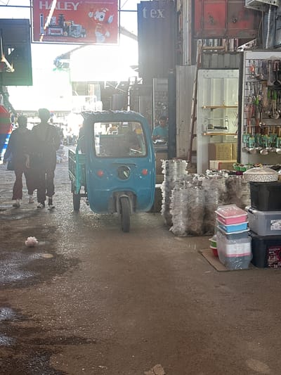 The image depicts a market scene in Kara-Suu, Kyrgyzstan. In the foreground, a bright blue three-wheeled vehicle, likely a tuk-tuk, is parked. To the left, two individuals, a male and a female, are walking away from the camera. The ground appears wet and reflective, suggesting recent rain or cleaning. In the background, various market stalls are visible, displaying an assortment of goods, including kitchenware such as pots, pans, and containers. A large billboard with the HALEY logo is prominently displayed at the top left, advertising kitchen appliances. The overall environment is consistent with a bustling outdoor market.