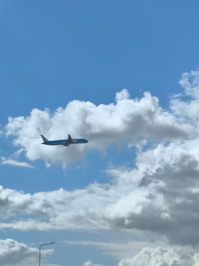 An airplane is flying in a partly cloudy sky. A light pole is visible in the bottom left corner. The location is likely Hoofddorp, Netherlands due to the KLM plane.