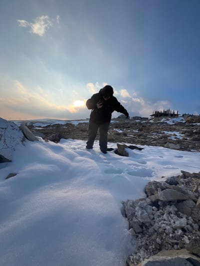 The image depicts an outdoor scene in Talfita, Syria, characterized by an uneven, snow-dusted, and rocky landscape. A solitary individual stands slightly right of center, positioned on an area where fresh snow meets exposed light-colored rocks and debris. The person is wearing a dark hooded jacket and dark trousers, and is hunched forward, holding a dark rectangular object, consistent with a smartphone, with both hands, appearing to focus on its screen.
The ground surface consists of irregular patches of pristine white snow intermingled with exposed light-colored rock formations and loose rubble. Footprints are faintly visible in some snow patches. In the middle ground, on an elevated mound to the right, stands an unfinished concrete building structure, showing exposed rebar and skeletal framework. The background horizon presents undulating terrain with some indistinct structures. The sky is partly cloudy, with the sun positioned low on the horizon to the left, creating a strong backlight and glare across the scene. The lighting suggests either early morning or late afternoon.
