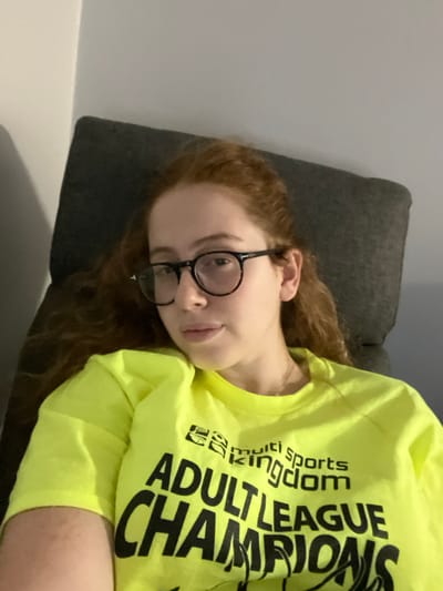 A young woman with reddish-brown, wavy hair and dark-rimmed circular glasses reclines against a dark gray upholstered chair or sofa. She wears a neon yellow short-sleeved t-shirt featuring black text that reads "multi sports kingdom" above "ADULT LEAGUE CHAMPIONS." Her face is visible from the forehead down, with a neutral expression and a slight pout. A plain, light-colored wall forms the background, showing a room corner on the left. The scene is indoors in Plainsboro Township, United States, under ambient lighting.