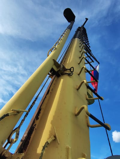 A low-angle shot showcases the mast of a ship, possibly a naval vessel, against a backdrop of a blue sky with scattered white clouds. The mast is constructed from yellow metal, with a prominent ladder on one side painted in black, extending upwards. A Russian flag is attached to the ladder. A cylindrical metal object, possibly a sensor or navigation aid, tops the mast. Ropes and chains are visible. The location is likely Gorushka, Russia, implied by the presence of the Russian flag on the mast.