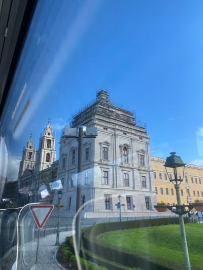 The image presents a daytime view of the Royal Palace and Convent of Mafra, Portugal, observed from inside a vehicle. The primary subject is a large, multi-story stone building with classical architectural features, predominantly grey in color. The central and upper sections of this building are covered in extensive scaffolding, indicating ongoing renovation or construction work. Several individuals, likely construction workers, are discernible on the scaffolding. To the left, two ornate bell towers with red-tiled roofs, part of the Basilica, rise prominently, featuring visible bells within their arches.
Adjacent to the main stone structure, on the right, a long, lower yellow/ochre building with numerous windows extends into the background. The immediate foreground features a manicured green lawn bordered by a neatly trimmed hedge, curving along a cobbled street. An ornate, dark green street lamp post stands in the mid-right foreground, and a modern lamppost is visible in the mid-left. A red and white inverted triangle "Give Way" traffic sign is prominently reflected on the vehicle's window in the lower left. The sky is clear and bright blue, with minimal cloud cover. Reflections on the window surface include streaks of light and distorted elements from the vehicle's interior.