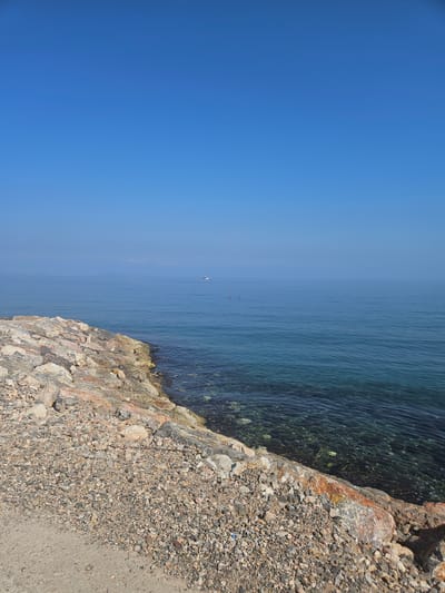 This image depicts a coastal scene in Güzelbahçe, Turkey, on a clear, bright day. The sky, occupying approximately the upper two-fifths of the frame, is a uniform, cloudless light blue, transitioning to a slightly paler hue near the horizon due to atmospheric haze.

The calm sea extends from the horizon to the foreground, exhibiting varying shades of blue and teal. In the mid-distance, near the center-left, two small, dark human figures are visible, appearing to be floating or swimming in the placid water. Further to the right and closer to the horizon, a small, white motorized boat or yacht is discernible as a distant speck. The sea surface is smooth with minimal ripples, reflecting the sky.

In the immediate foreground, the water transitions to a clear, greenish-blue, revealing the rocky seabed below. Submerged rocks and what appears to be dark aquatic vegetation are visible through the transparent water.

The shoreline on the left and bottom of the image consists of a rocky embankment or breakwater. This structure is composed of irregularly shaped rocks of varying sizes, exhibiting a spectrum of colors including gray, white, brown, and reddish-brown, with some patches of yellow or orange discoloration, possibly from algae or mineral deposits. This rocky edge descends into the clear water.

Adjacent to the rocky embankment, the foreground land consists primarily of coarse gravel and small pebbles, displaying a mixed coloration of grays, browns, and whites. In the lower-left corner, a smoother, lighter-colored dirt or compacted earth path transitions into this gravelly terrain. Minor anthropogenic debris, such as a small blue plastic fragment and a stick, are interspersed among the gravel. The overall scene suggests a tranquil, unpopulated coastal environment.