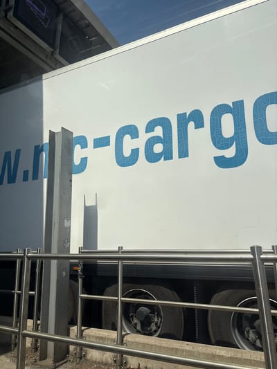 The image shows the side of a white truck with blue lettering that reads "w.n.c-cargo". The lettering is large and stylized, with a textured pattern within the blue. The truck is parked outdoors, with a clear blue sky visible above. In the foreground, there is a metal railing and a concrete curb. To the left of the truck, a vertical metal support is visible, casting a shadow on the truck's side. The scene appears to be in an industrial or commercial area. The location is identified as Le Boulou, France.