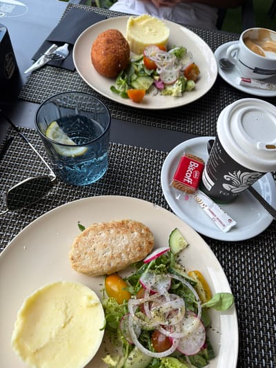 A table set for a meal outdoors. Two plates of food are visible: one with a salad and a breaded patty, and another with a fried ball, mashed potatoes, and a salad. A glass of water with lemon and a pair of sunglasses are on the table, along with a cup of coffee and a small cookie. The table is covered with a woven placemat. The scene appears to be in a restaurant in La Vila Joiosa, Spain.
