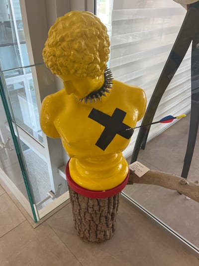 An upward-angled, eye-level shot captures a bright yellow, stylized bust of a male figure with curly hair, positioned in an indoor setting. The bust wears a black choker adorned with silver-colored spikes. A prominent black 'X' symbol is affixed to the center of its chest. An arrow, featuring a black shaft and red, blue, and yellow fletchings, is embedded into the bust's chest to the right of the 'X'. The bust rests on a red circular base, which in turn sits atop a cylindrical pedestal crafted from textured tree bark. The floor is a light-colored, possibly tiled, surface. To the left, a clear glass barrier reflects elements of the surrounding environment, including a white structural beam. In the background, a window with horizontal white blinds is visible, and to the right, a dark, angular display support is partially present. A light-colored wooden branch with an identification label, partially obscuring the text "LINO PALADETTI", extends from the right foreground towards the base of the pedestal. This scene is located in the city of Galatina, Italy.