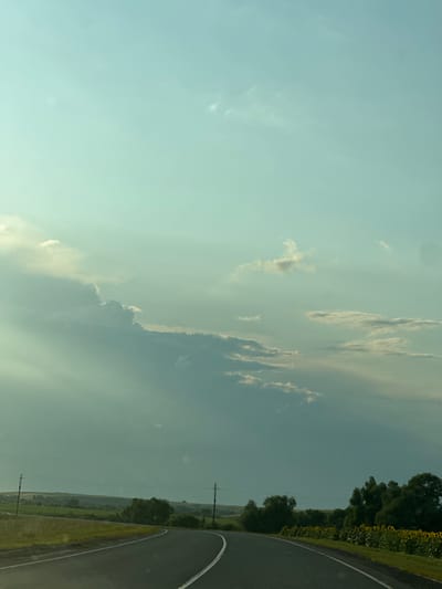 The image captures a rural road curving through a landscape under a sky with scattered clouds and sunbeams. The road itself is asphalt with white lane markings. To the left of the road, there's a grassy field bordered by more fields and distant rolling hills. Power lines with poles are visible along the roadside. To the right, a dense line of trees stands, and beyond them, a field of sunflowers is in bloom. The overall impression is one of a scenic drive in a rural area.