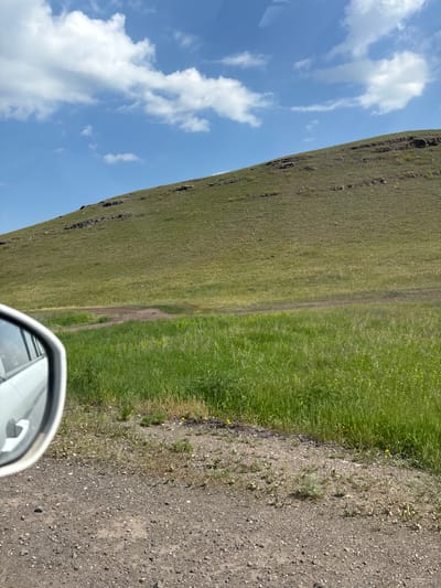 The image depicts a rural landscape with a grassy hill under a partly cloudy blue sky. In the foreground, a gravelly dirt road is visible, and on the left side, a portion of a white car's side mirror is reflected. A patch of lighter-colored dirt is present in the middle ground, contrasting with the surrounding green grass. The hill itself appears uneven, with some rocky outcrops and sparse vegetation. The overall scene suggests a natural, undeveloped area, likely outside of a populated urban center. The question of its location within the city of Borodino, Russia, cannot be answered from the visual information provided.