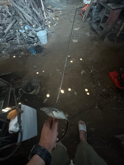 The image depicts an indoor scene, possibly a workshop or storage area, with a rough, dirt floor littered with debris. In the foreground, a person's right hand is visible, holding a metallic object with a curved edge, possibly a shield or part of a tool. The person is wearing a dark watch on their left wrist and is clad in olive-green pants. A light-colored sandal is on their right foot. Scattered throughout the scene are numerous small, circular yellow lights on the floor, their source unclear but suggesting some form of illumination or perhaps debris reflecting light.
To the left, a large pile of metal scrap, including pipes and various irregular metal pieces, is stacked. A grey metal bucket and a smaller black bucket are near this pile, along with a blue object, possibly another bucket or container. Further into the scene, a red object, which appears to be a piece of equipment or machinery, is visible on the right. A long, thin pole extends from the foreground towards the back of the space. Shelving units containing various items are also present in the background. The overall environment appears somewhat cluttered and industrial. The specific mention of Terny, Ukraine, is an external piece of information not directly derivable from the visual content of the image itself.