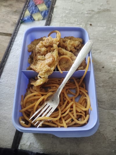 The image shows a light blue rectangular lunchbox containing two separate compartments of food. The left compartment holds a portion of fried chicken or other crispy fried food, which appears golden brown and textured. The right compartment contains a pile of spaghetti, which is also golden brown and appears to be lightly sauced or seasoned. A silver fork with a decorative pattern on the handle rests across both compartments, with its tines inserted into the spaghetti.
The lunchbox is placed on a textured surface that appears to be a table or placemat. In the background, to the left of the lunchbox, a corner of a patterned fabric or mat is visible, featuring geometric shapes in red, blue, and yellow. The overall lighting suggests it might be daytime.
The scene depicts a meal, likely ready to be eaten. The food appears to be a casual, home-style meal. There is no indication of any people or specific activities beyond the preparation and presentation of the food. The setting is simple and functional, with the focus primarily on the lunchbox and its contents. No visible text is present in the image. The location context of Cintadamai, Indonesia, implies a domestic or casual dining setting within that region.