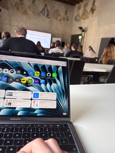 A first-person perspective photograph captures a person operating a silver MacBook Pro laptop on a white table within a lecture or conference room in Krems an der Donau, Austria. The laptop screen displays a web browser with "Google Gemini" in the search bar, a tab labeled "Barbershop Idea - Google Docs," and a start page featuring a grid of application icons (e.g., AI Fluent, Motion, NVIDIA Developer) and recent search history synced from an iPhone (e.g., "Round trip to Berlin," "Balea Med Ultra Sensitive Intensivcreme"). A human hand is positioned over the QWERTZ keyboard. In the background, approximately ten to fifteen individuals are seated in rows, mostly seen from behind, observing a large projection screen displaying a presentation with partially visible white text on a light background (e.g., "Google I"). A male wearing a dark suit jacket is prominently seated with his back to the viewer in the immediate foreground of the background. The room features light-colored walls adorned with faded historical murals or frescoes, dark chairs, light tables, and visible wooden ceiling beams, suggesting an older, repurposed building.