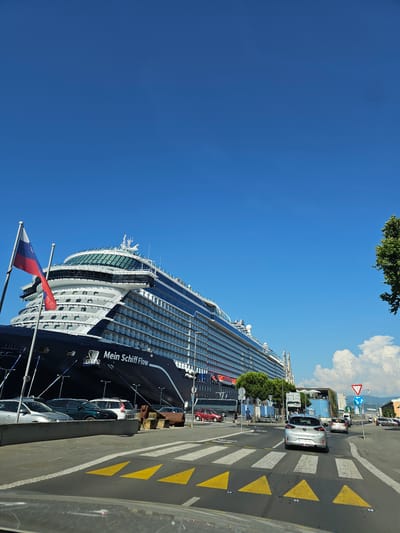 This image depicts a street scene in the city of Koper / Capodistria, Slovenia, with a large cruise ship docked in the background. The cruise ship, identified by the text "Mein Schiff Flow" on its hull, is dark blue and grey, with numerous decks and windows. It is moored at a port, with mooring lines visible. In the foreground, there is a paved street with a pedestrian crossing marked by white stripes and yellow triangular directional arrows. Several cars are present on the street, including a silver hatchback in the center of the frame, a red sedan to the left, and a parked silver sedan and other vehicles to the left of the cruise ship. A Russian flag is flying on a flagpole to the left of the ship. The sky is clear and blue, with some white clouds visible in the distance. Trees and buildings are also present in the background, suggesting an urban port environment. The scene appears to be a sunny day, with good visibility.