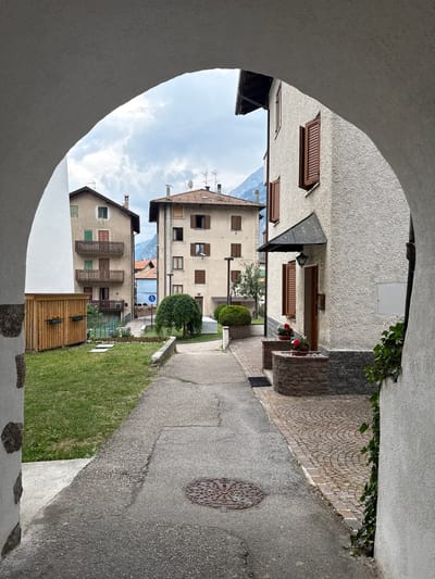 This is a medium shot of a quaint Italian village street, likely Molveno, Italy, as indicated by the architecture. The image is captured from within an archway, framing a view of a narrow, paved street lined with traditional buildings. The buildings are multi-story with stone and stucco facades, featuring wooden shutters on many windows, suggesting a European aesthetic. Ivy climbs up some of the walls, adding a touch of nature to the urban setting.

The scene appears to be during the daytime, under a cloudy sky. There are no people visible in the image, contributing to a sense of quiet serenity. The street is bordered by a grassy area on one side and a cobblestone path on the other, leading towards the background where more buildings and hints of mountains are visible. A circular manhole cover with an intricate pattern is a notable detail on the paved street. Flower pots with red flowers are placed near the entrance of one of the buildings, adding a splash of color. The overall mood is peaceful and evocative of a small, historic European town. No visible text is present in the image.