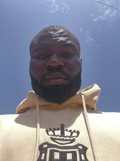 A low-angle, tilted, outdoor close-up photograph shows a dark-skinned man from the chest up against a pale blue sky, likely in the city of Mbar, Nigeria. His head is angled towards the left side of the frame. He has short, dark hair and a full, dark beard. The man wears a light tan or cream-colored hoodie with a black graphic design on the front. The hood is up. The hoodie has drawstrings that are light tan or cream-colored and have white or light-colored tips. The man is looking slightly down at the camera, and his facial expression is neutral. The sky is a clear, pale blue, suggesting a sunny day. A cable is visible on the left upper corner.
