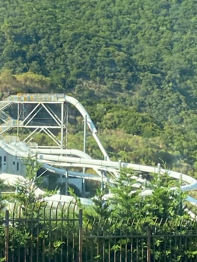 The image shows a water slide complex in Seoce, Montenegro, nestled against a backdrop of lush green trees. The water slide is made of white plastic tubing and is supported by a metal structure. There is a building visible at the bottom of the slide. A black metal fence runs across the foreground, partially obscuring the view of the water slide. The trees in the background are dense and cover a hillside. The lighting suggests it is daytime.