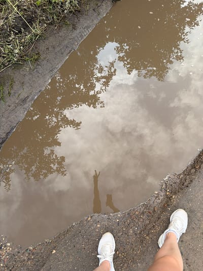 This image shows a person's legs and feet wearing white athletic shoes, standing at the edge of a muddy puddle. The puddle reflects the sky, clouds, and tree branches. The ground around the puddle is a mix of mud, gravel, and some dry grass. The scene appears to be outdoors, possibly on a dirt road or path. There is no specific indication of the location being in the city of Upeslejas, Latvia.