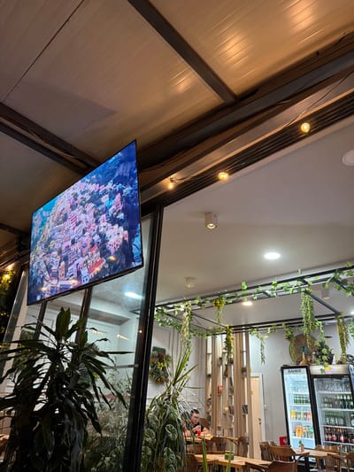 The image captures the interior of a cafe or restaurant. A large flat-screen television is mounted on the wall, displaying a vibrant aerial view of a coastal town with colorful buildings. The ceiling features a corrugated metal structure with exposed beams and decorative string lights. Lush green plants are strategically placed throughout the establishment, with some hanging from the ceiling and others in large pots. A glass-fronted refrigerator stocked with beverages is visible on the right. In the background, a person is seated at a table, appearing to be eating. The overall ambiance suggests a casual and well-decorated space. The city of Krumovgrad, Bulgaria is mentioned in the prompt, but there is no visual information in the image to confirm this specific location.