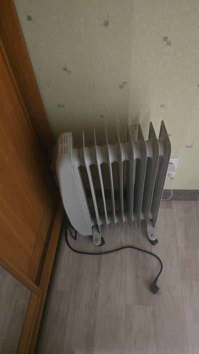 The image shows a white electric oil-filled radiator placed on a light wood-grain laminate floor next to a wooden cabinet or door. The radiator has multiple fins and a control panel on the side. A black power cord extends from the radiator to the right. In the background, the wall is covered with a light-colored wallpaper with a subtle pattern. A white electrical outlet is visible on the wall behind the radiator. The scene appears to be indoors, likely in a residential setting. There are no people or animals present. The lighting suggests it is daytime. The location is identified as Ukhta, Russia.