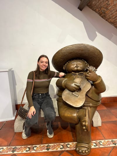 A young woman poses beside a bronze statue of a man playing a guitar. The statue is wearing a large sombrero and appears to be seated. The woman wears a long-sleeved olive green shirt with stripes, gray jeans, and sneakers. She holds one arm up in a playful gesture, appearing to interact with the statue. A small purse hangs from her shoulder. The background features a white wall, a tiled floor with decorative patterns, and partial views of architectural details.