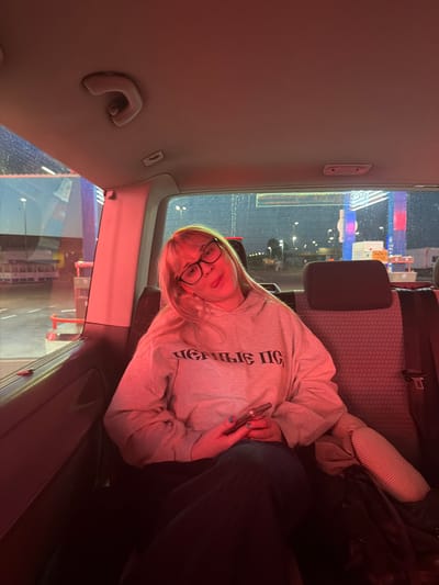 The image depicts a woman in a car. She is wearing glasses and a grey sweatshirt, holding a phone. The background seen through the car windows suggests a nighttime scene, possibly a fair or amusement park, with bright lights and structures. The presence of rain on the windows is also visible. The reflection in the windows show signs that they are at the Pazova Fair, Stara Pazova Municipality, Serbia.